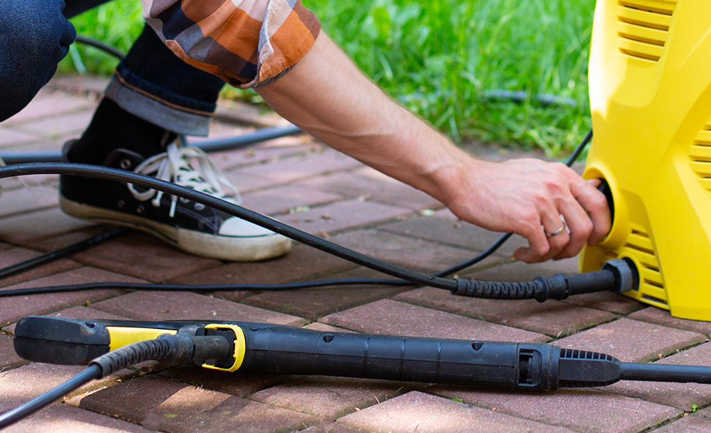 A person wearing black sneakers and rolled up jeans squats down to attach a hose to a yellow pressure washer.