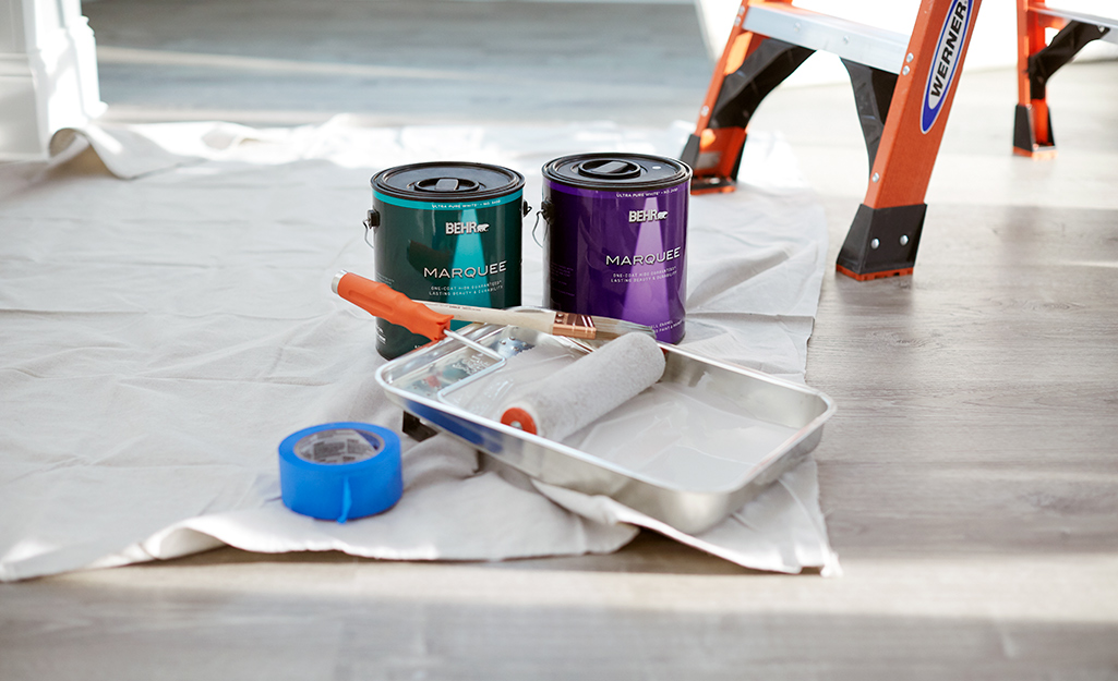 Paint primer, painter's tape, a paint roller and paint tray sit on a drop cloth on a floor.