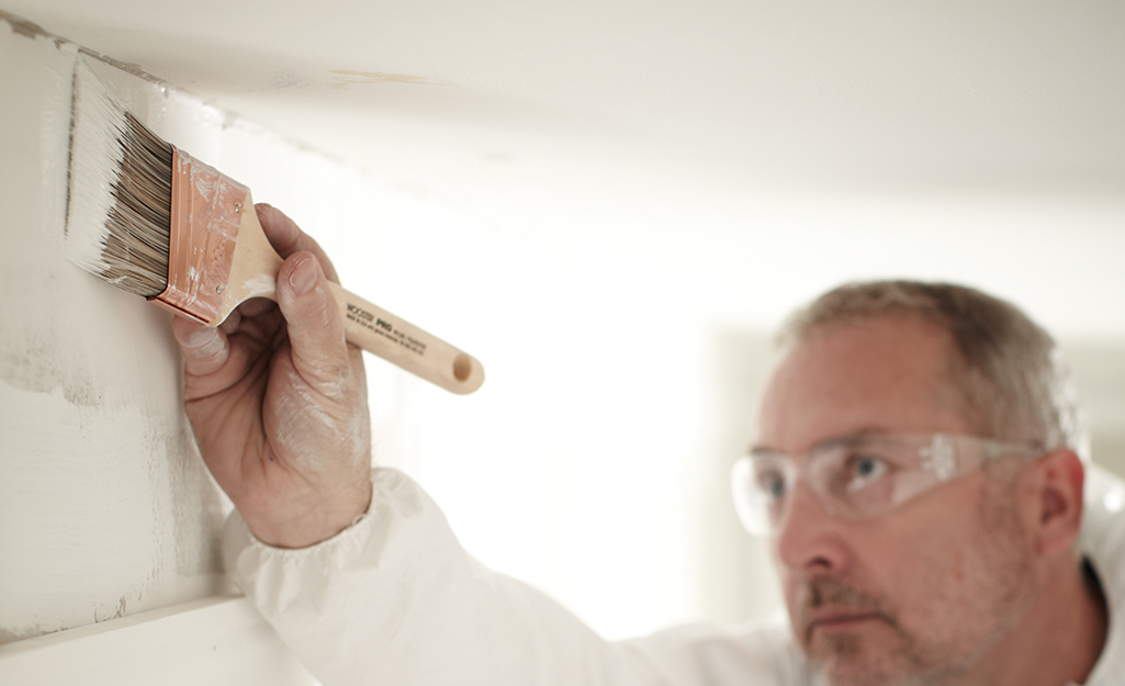 A person using a paint brush to apply primer on a wall at the edge of the ceiling.