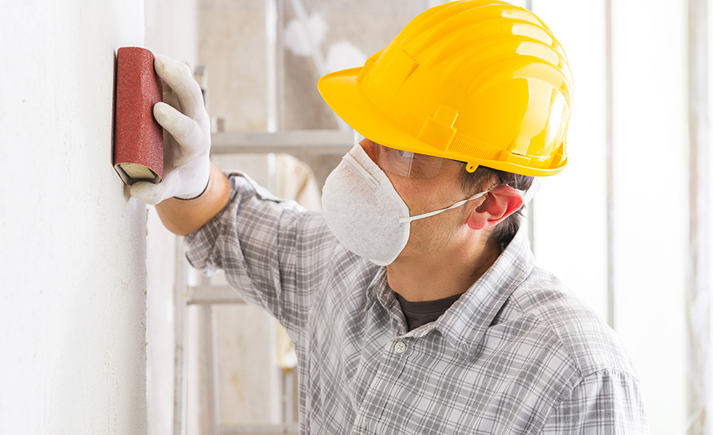 A person wears a dust mask while using a sanding block to smooth a coat of primer on a wall.