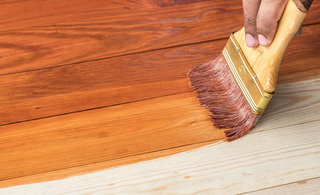 A person applies wood stain to a hardwood floor with a brush.