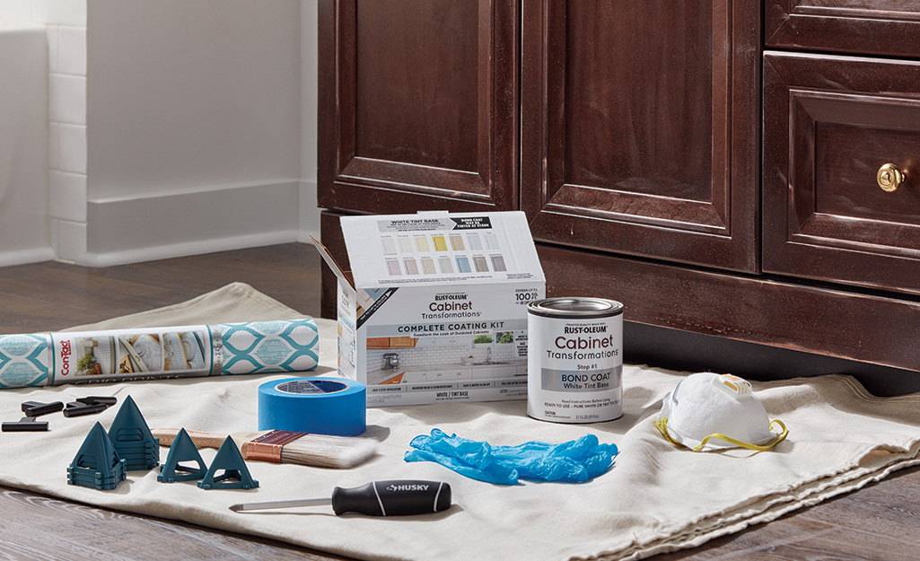 A screwdriver, gloves, paint, a paint brush, contact paper and cabinet hardware lay on a drop cloth in front of a bathroom vanity.