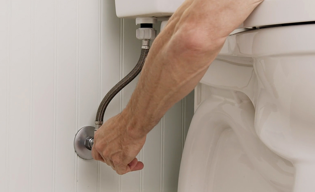 A person turning off the water shutoff valve behind a toilet.