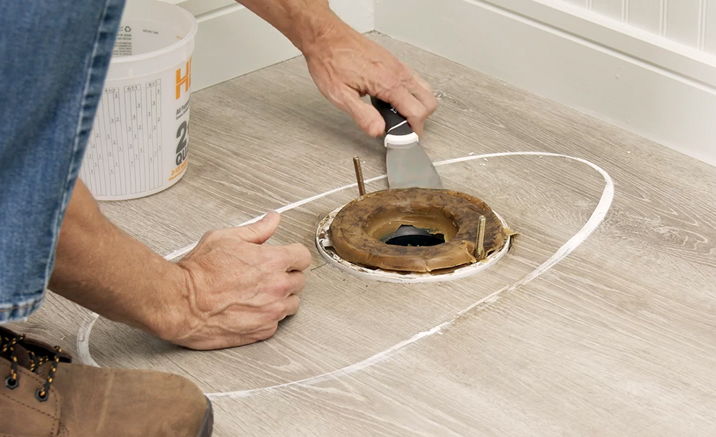 A person using a paint scraper to remove an old toilet wax ring from the toilet flange.