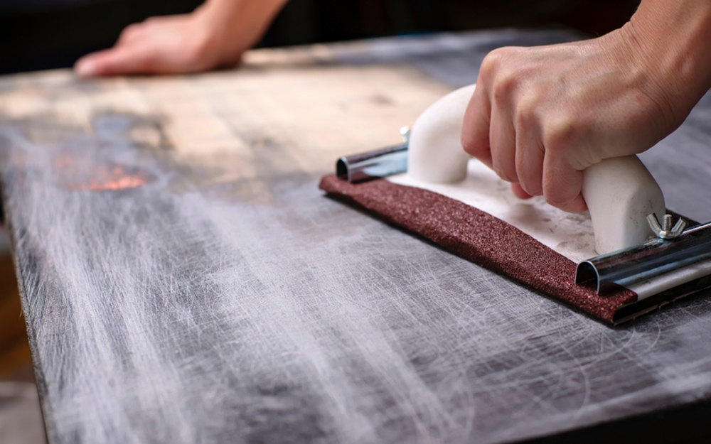 A person using a manual hand sander and sandpaper to remove paint from wood.