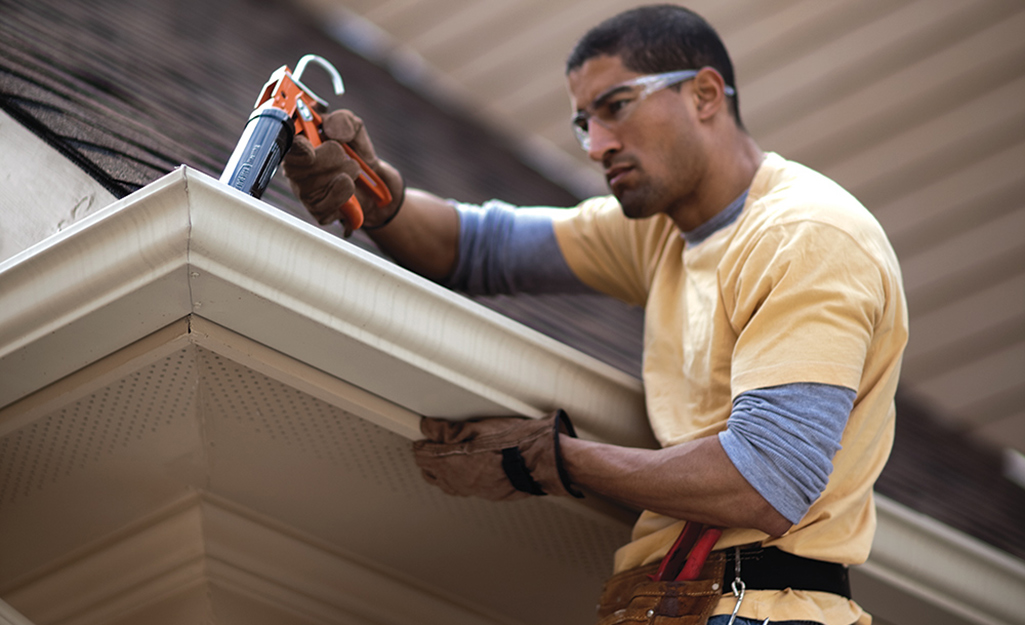 A person adding plastic roofing cement around white gutters on a roof's edge.