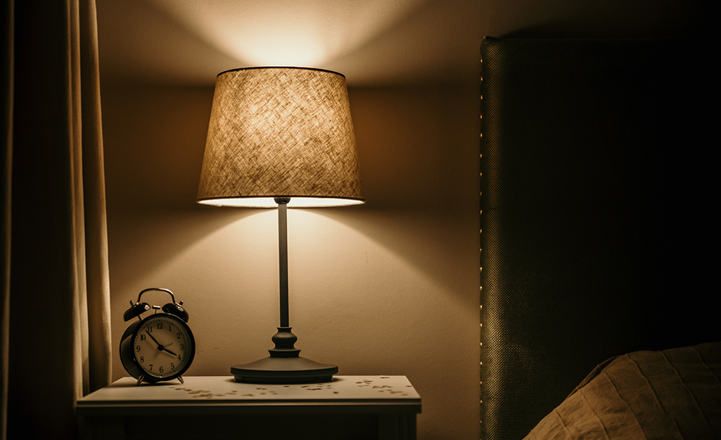 A rewired lamp on a bedside table illuminates a vintage alarm clock and bed.