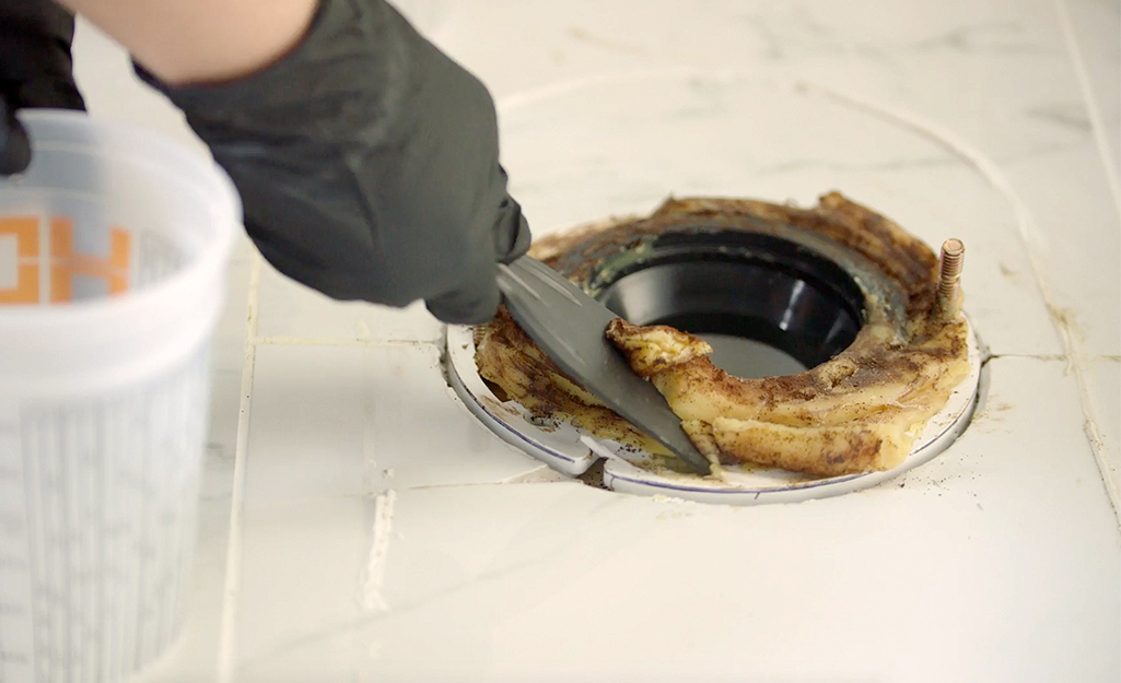 A person using a paint scraper to remove an old wax ring on a toilet flange.
