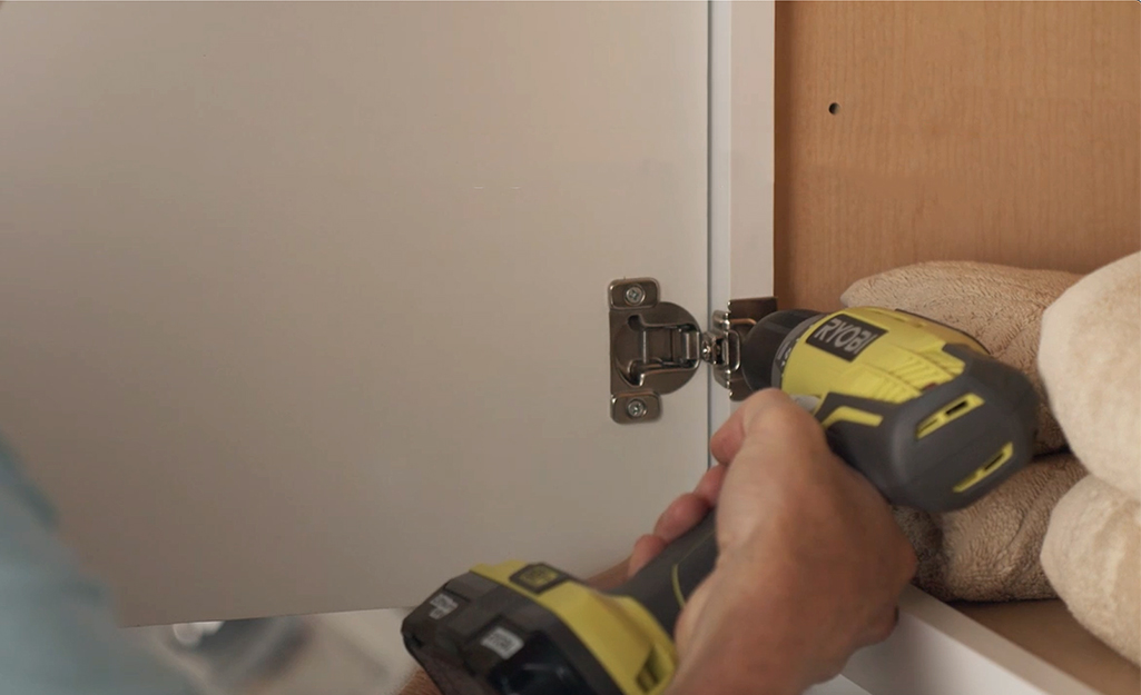 A person removing an old hinge from a kitchen cabinet door.