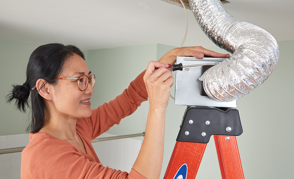A woman standing on a ladder removes an old bathroom exhaust fan from its ductwork.