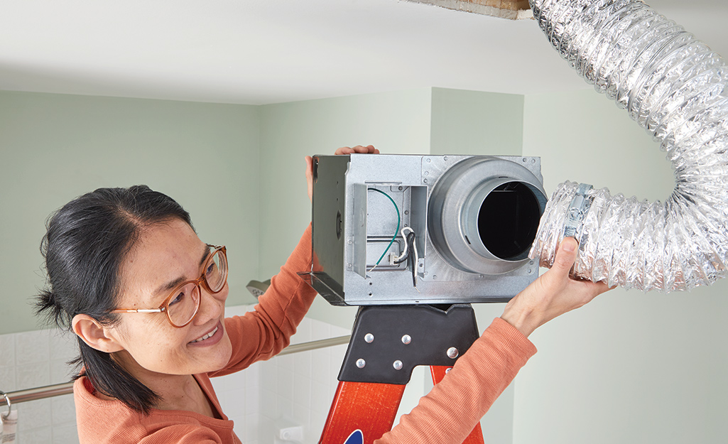 A woman standing on a ladder attaches a new bathroom exhaust fan to a duct.