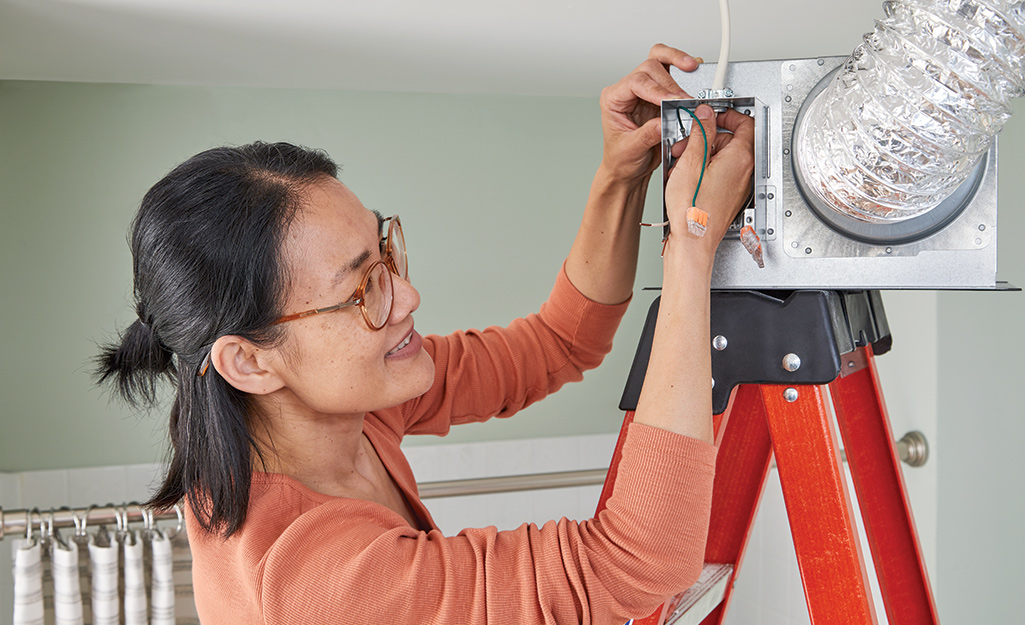 A woman standing on a ladder connects the wiring for a new bathroom exhaust fan.