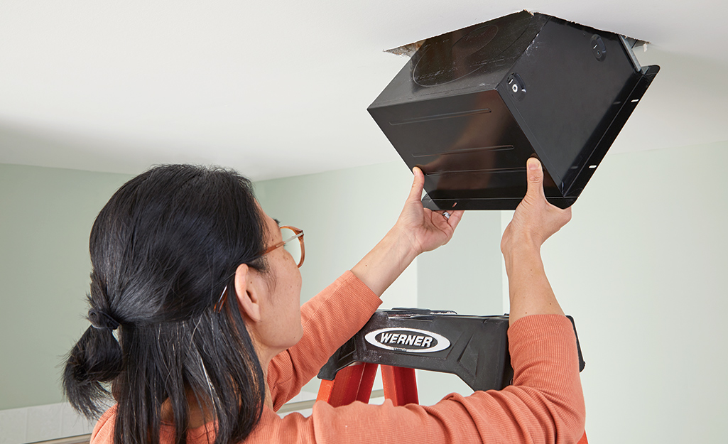 A woman places a bathroom exhaust fan into its housing in the ceiling.