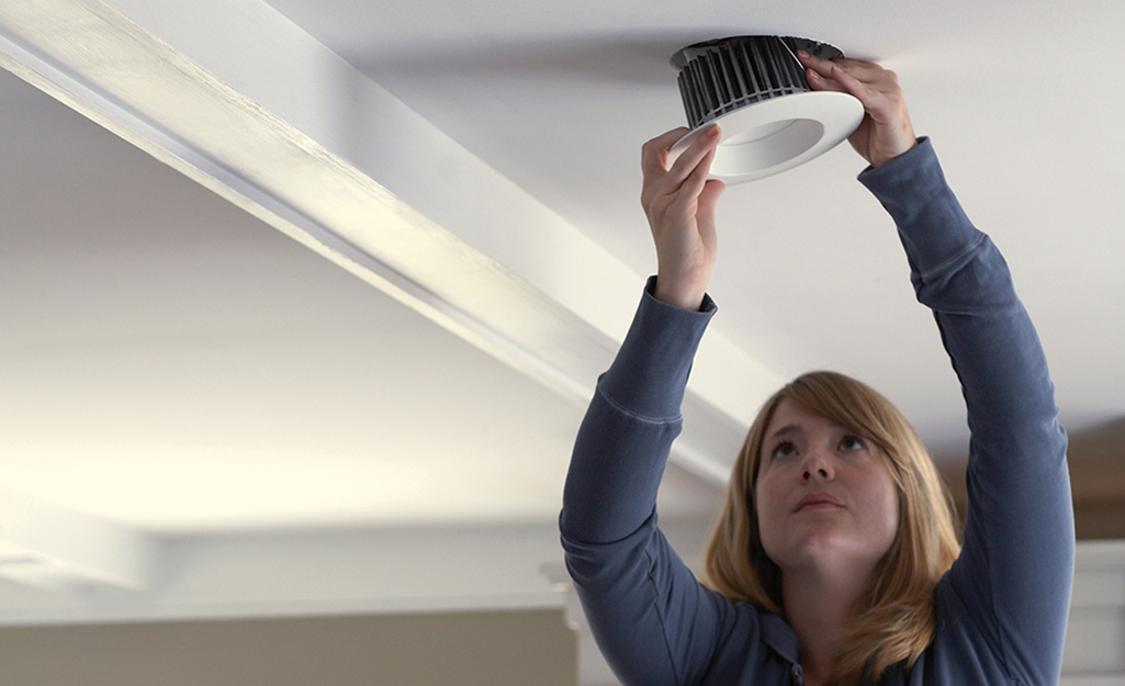 A woman reaches up to take a recessed lighting fixture out of the ceiling.