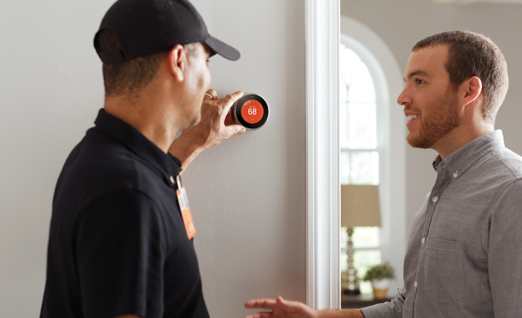 A Home Depot associate standing next to a homeowner and adjusting a smart thermostat on a wall.