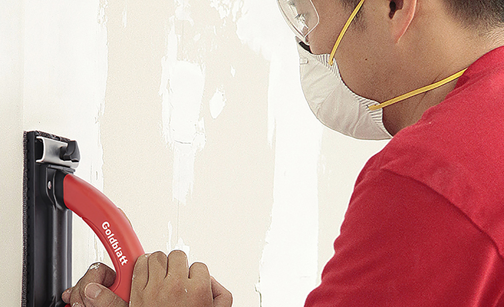 A person wearing safety glasses and a mask sands a section of drywall.