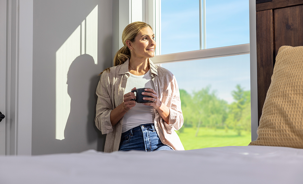 A woman holding a coffee mug sits leans against a window sill in a bedroom.