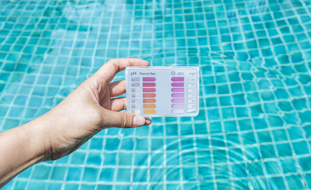 A hand holds a pH test above the clear water of a swimming pool. The bottom of the pool is visible.