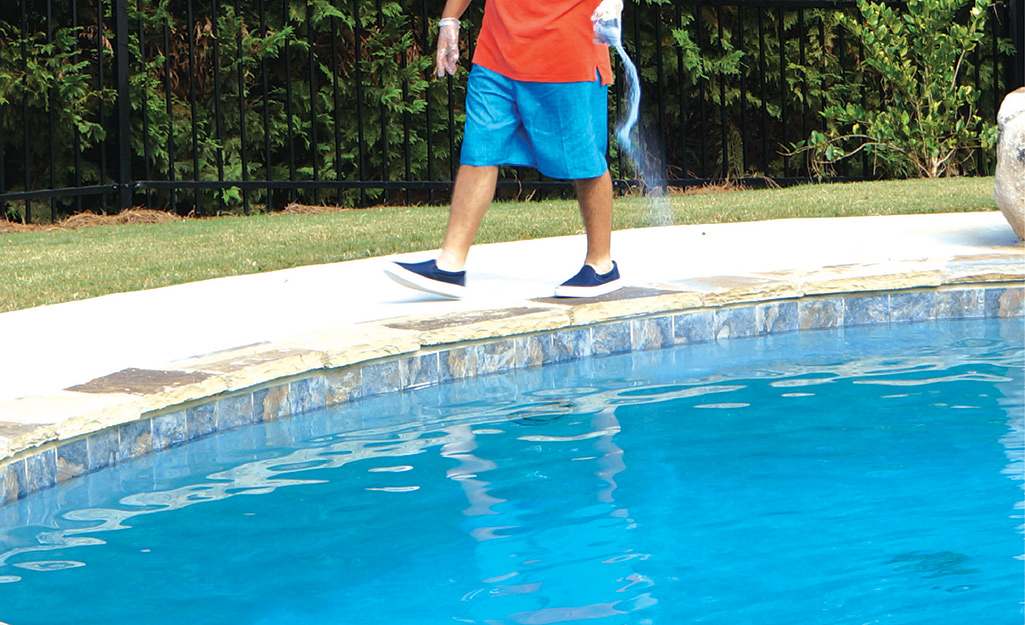 A man shakes a package of granular pool shock into the blue water of a swimming pool as he walks around it.