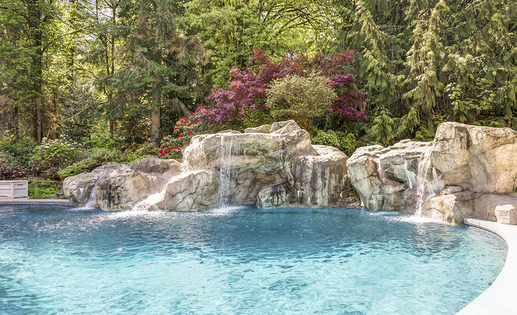 Waterfalls flow down large stones into a swimming pool in a natural setting of trees and blooming plants.
