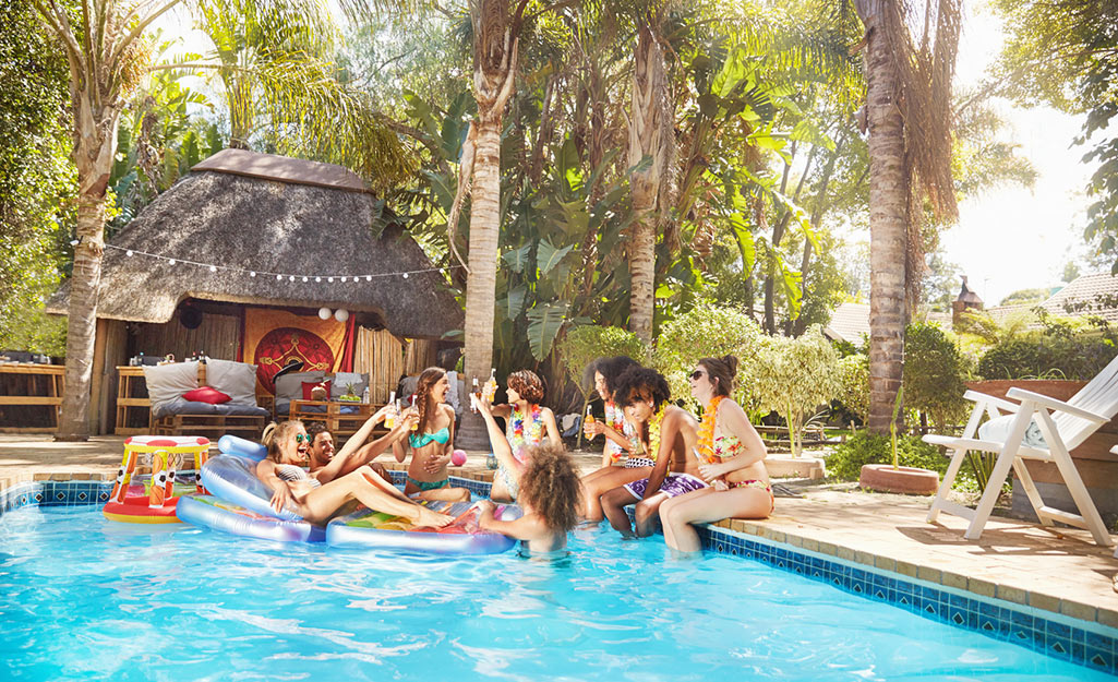 Eight people wearing swimsuits and floral leis hold bottled beverages and laugh while in a pool next to palm trees.