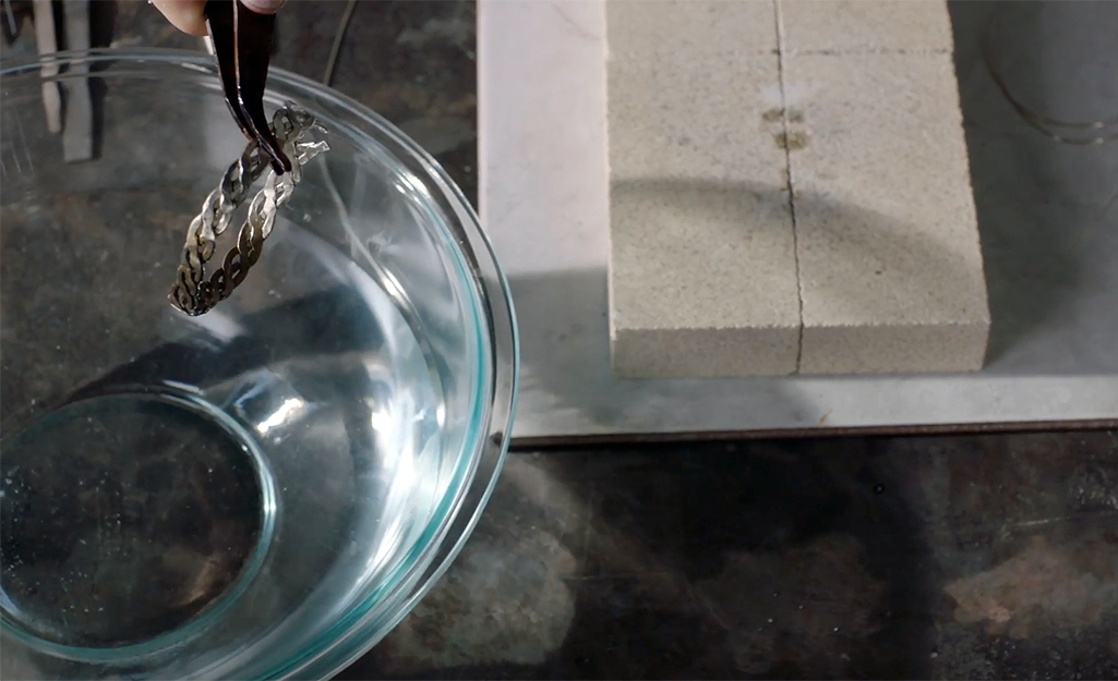 A person transfers a soldered bracelet to a bowl of water using pliers.