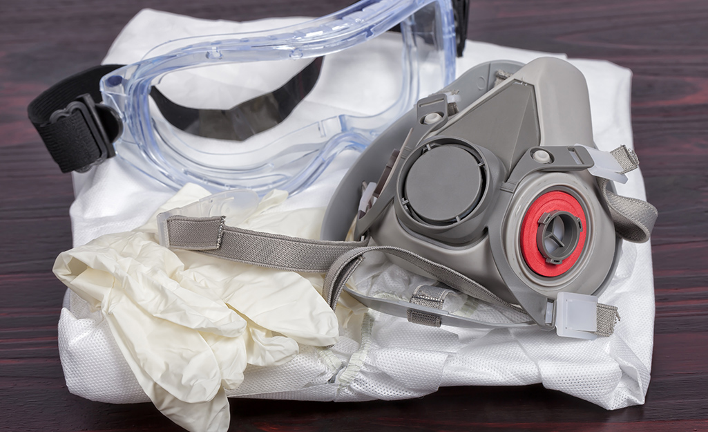 Safety goggles, gloves and a respirator sitting on top of folded white paint coveralls.