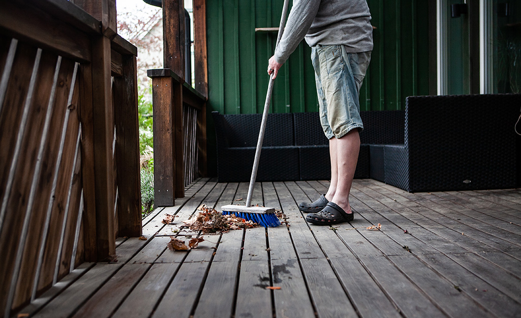 A person sweeps leaves off a deck with a push broom with blue bristles. 