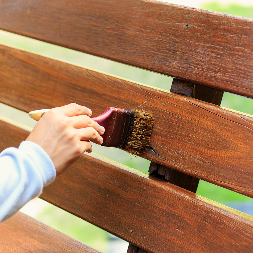 A person uses a brush to stain a bench made of pressure-treated wood.