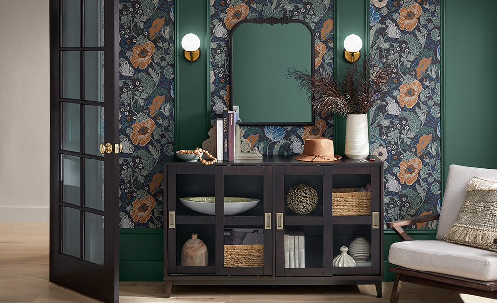 A buffet table paired with dark green painted walls, rich floral wallpaper, wall sconces and a modern side chair