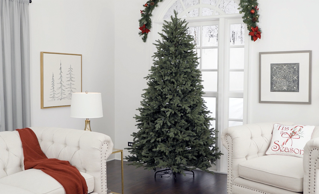 An artificial Christmas tree stands in front of window in a living room with a white sofa and a white chair.