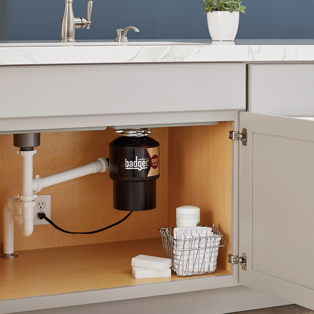 A cabinet under a kitchen sink stands open, showing pipes, a garbage disposal and a basket with towels and cleaning supplies. A cabinet under a kitchen sink stands open, showing pipes, a garbage disposal and a basket with towels and cleaning supplies.