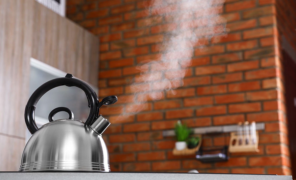 A silver kettle boils on a stove and its steam is visible against a brick wall.