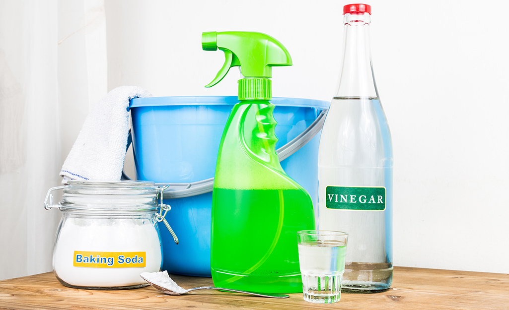 A container of baking soda sits next to a blue bucket, a green spray bottle and a bottle of vinegar.