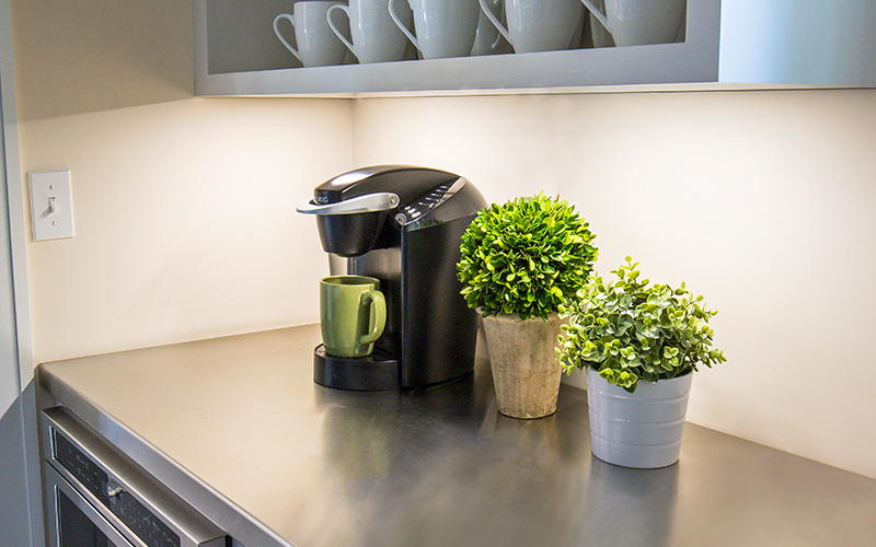 Under-cabinet lighting shines down on a coffee pot and plants.