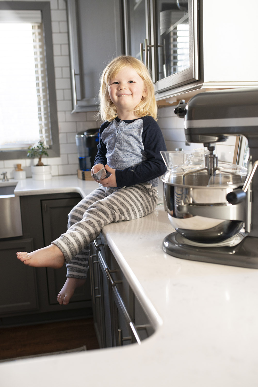 A young child sitting on a white kitchen countertop next to a KitchenAid stand mixer.