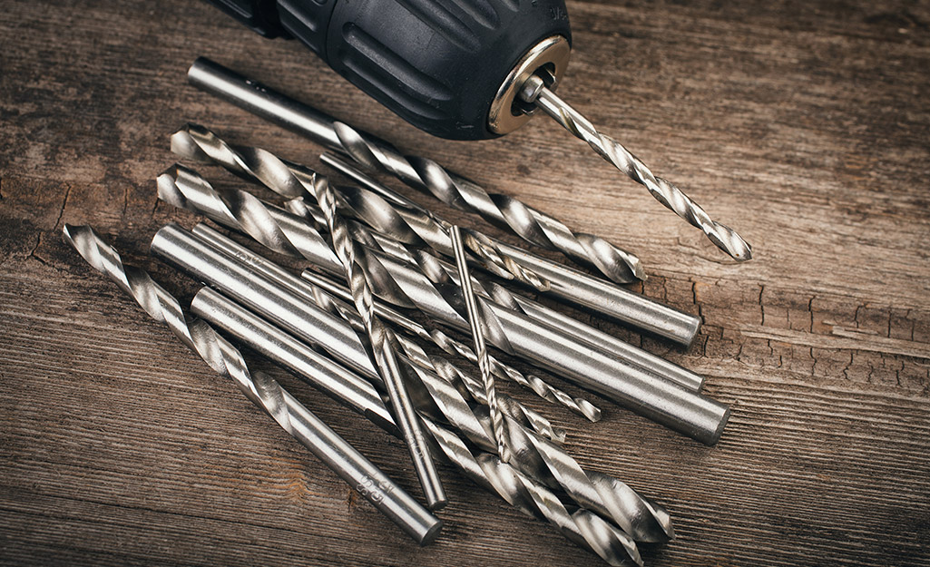 A pile of twist drill bits and a drill sit on a wooden table.
