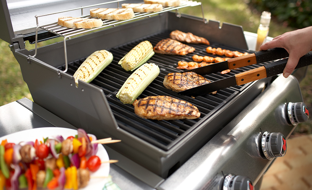 A person tending to a variety of food cooking on a gas grill.