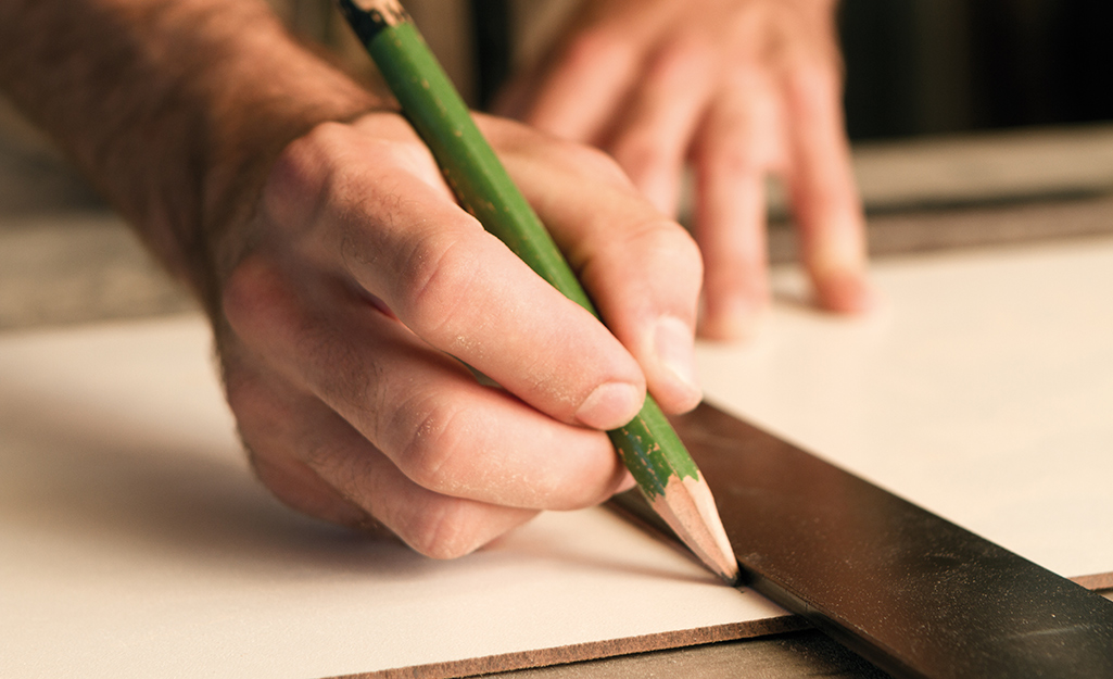 A person uses a pencil to mark a cutline on a piece of wood.