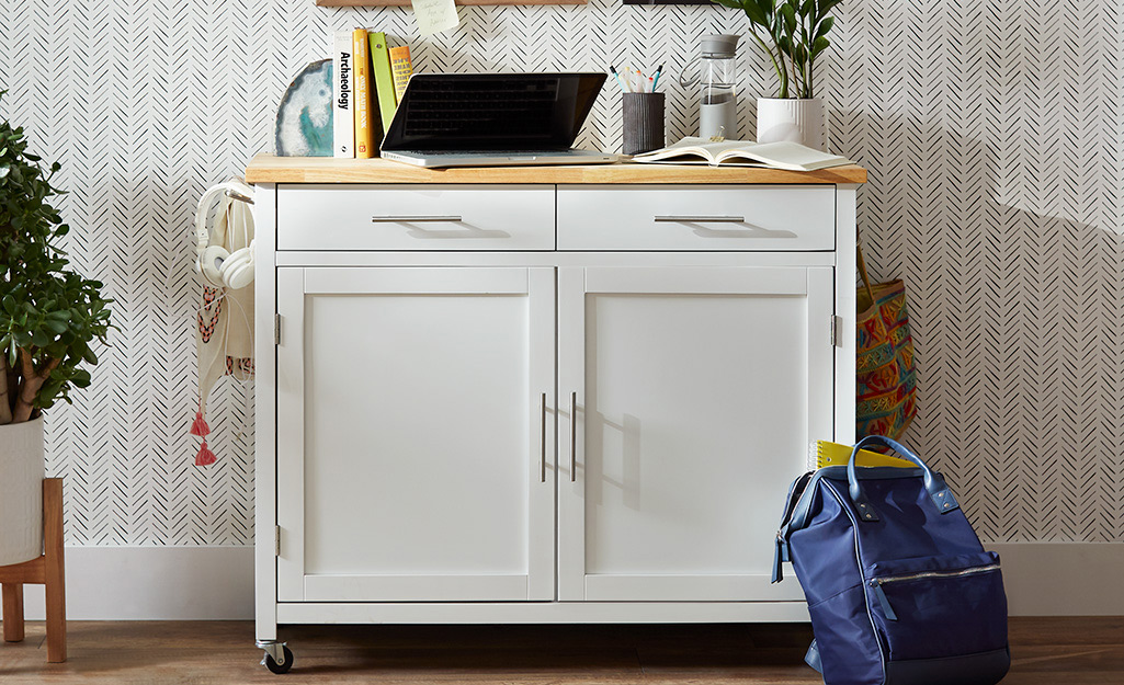 A kitchen cart used as a work station, with a laptop and books on its top and accessories hanging from its towel bar.