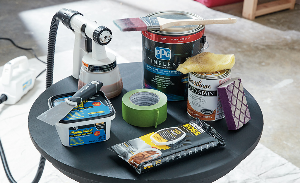 A paint sprayer, paint, wood stain, painter's tape, wood filler, cleaning clothes and a chisel sit on a small round table. 