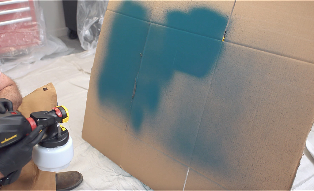 A person sprays blue paint on a piece of cardboard to practice using a paint sprayer.
