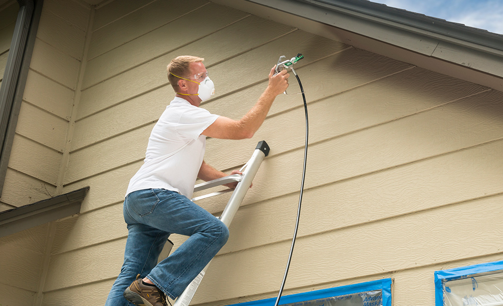 A man standing on a ladder uses a paint sprayer to paint the siding on a house.