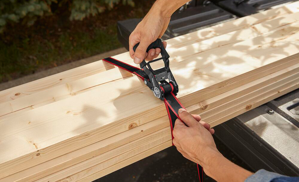 A person uses a ratchet strap to secure a small load of lumber.