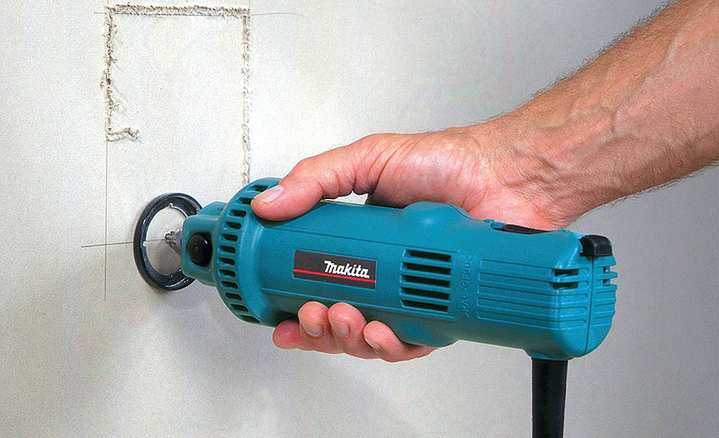 A large, corded rotary tool cuts a square out of drywall.