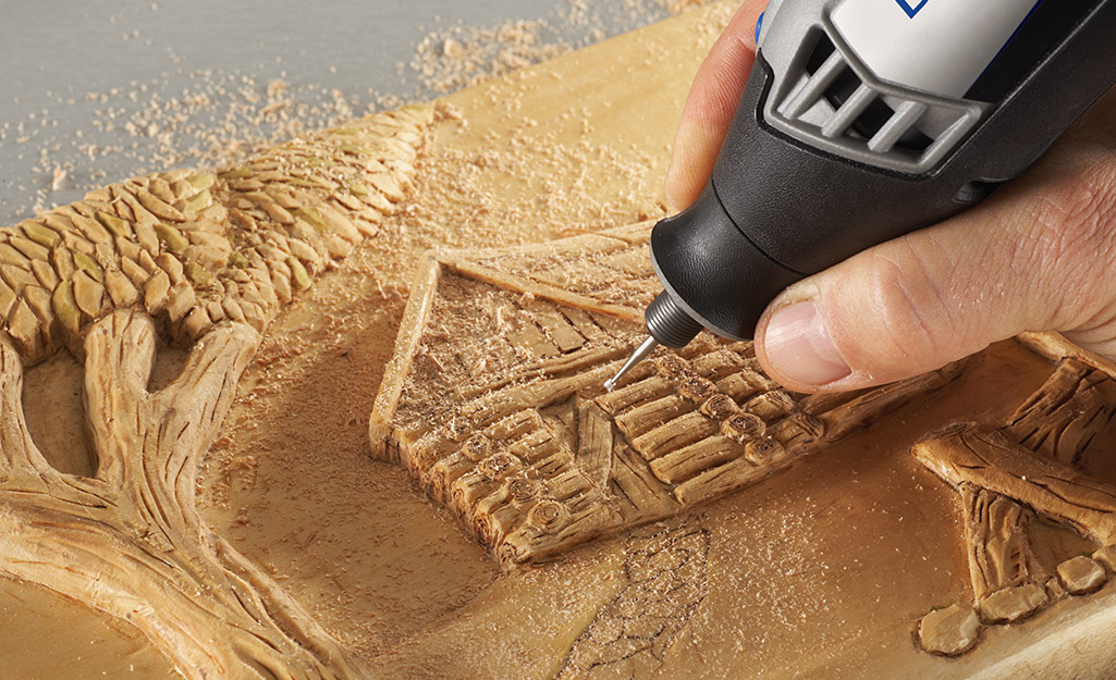 A person carves an illustration of a tree and cabin into a piece of wood using a rotary tool.
