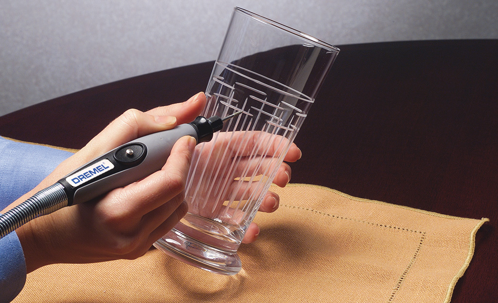 A person uses a rotary tool to etch a design into a drinking glass.