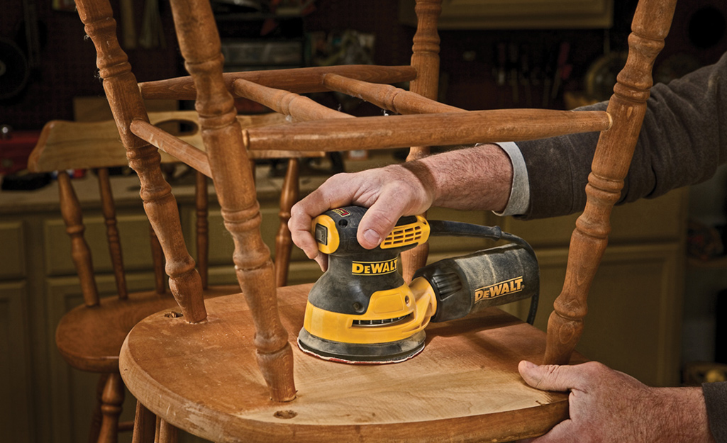 A person uses a random orbital sander to remove the finish from the bottom of a wood chair.