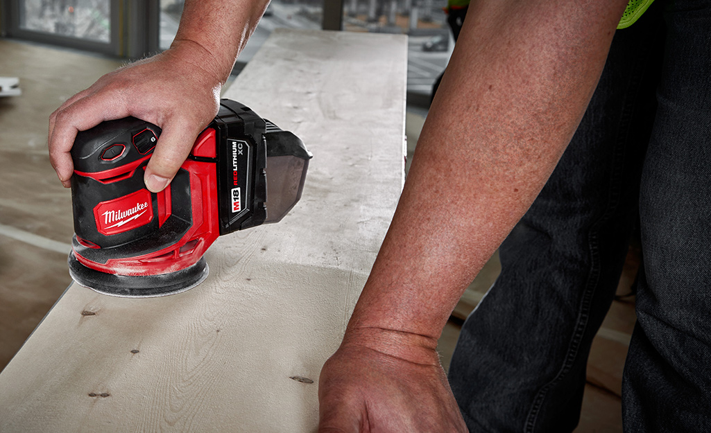 With one hand operating a random orbital sander, a person sands a piece of wood.