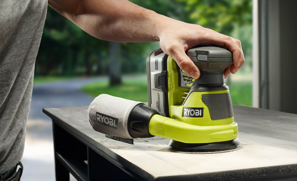 A person uses one hand to operate a random orbital sander as they sand a piece of furniture in a garage workshop.